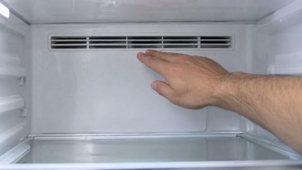 The rear wall of an empty refrigerator compartment with a narrow air vent grille and a hand held near the grille